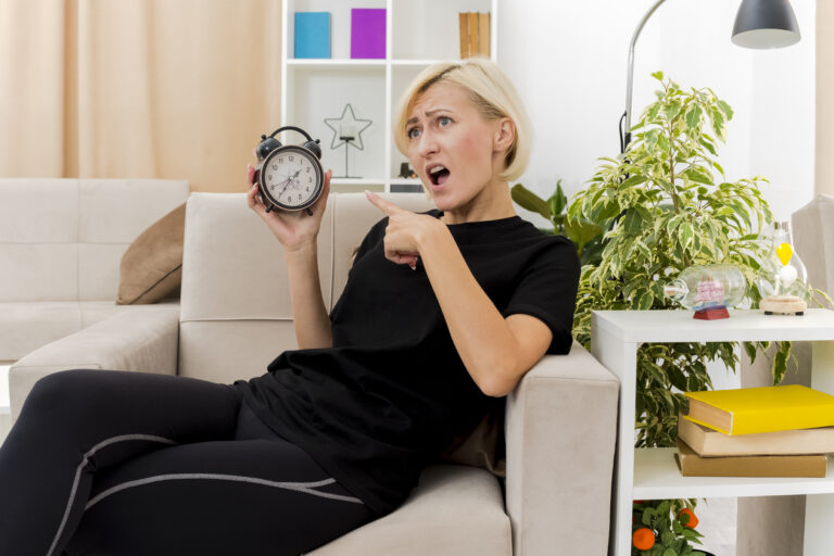 annoyed beautiful blonde russian woman lying on armchair holding and pointing at alarm clock inside design living room